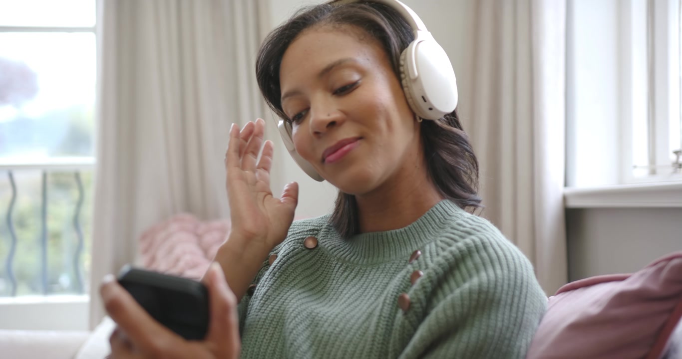 Donna afroamericana che regola le cuffie mentre usa il telefono sul divano vicino alla finestra, cambiando canzone.