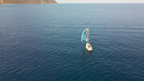 Velero-Con-Velas-Coloridas-Navegando-En-El-Mar-Azul-Hacia-La-Costa-En-El-Fondo-En-Cerdeña,-Italia---Toma-Aérea-De-Drones