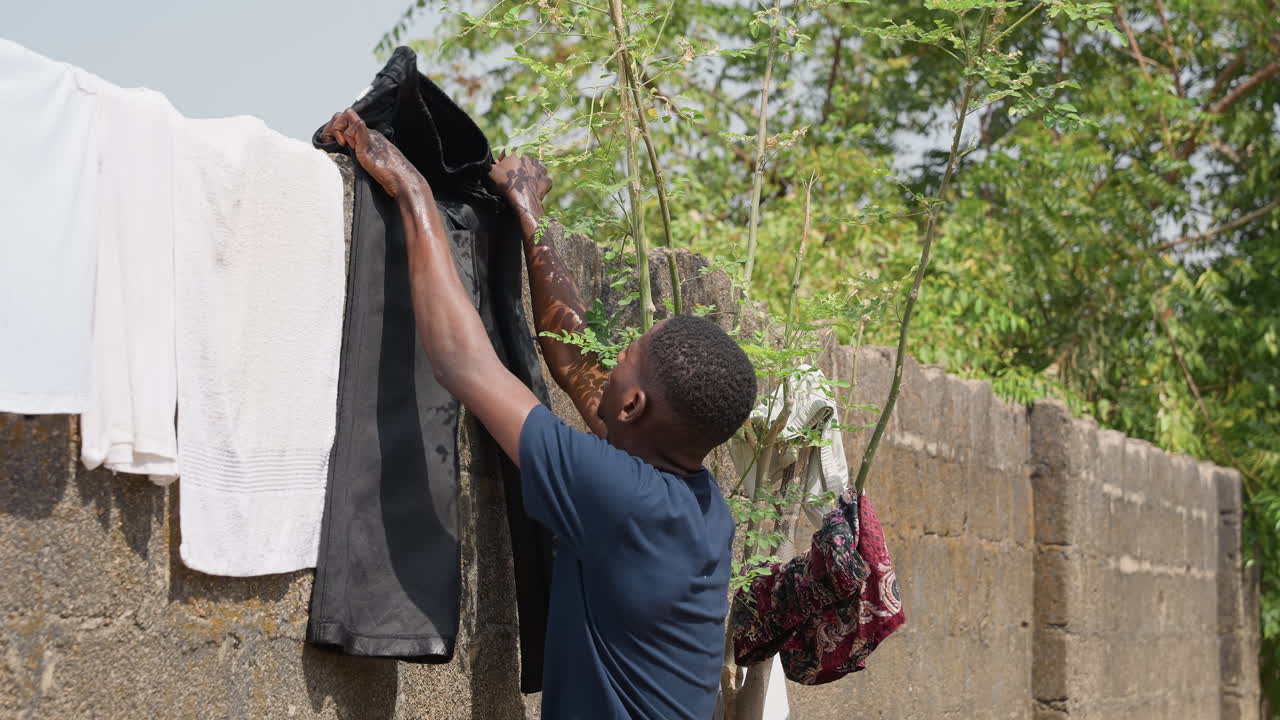 Teen hangs laundry. Youth drapes wet fabric outside. Teenager carefully hangs garments on house exterior. Young individual assists by placing damp clothing on wall surface for drying in sunlight