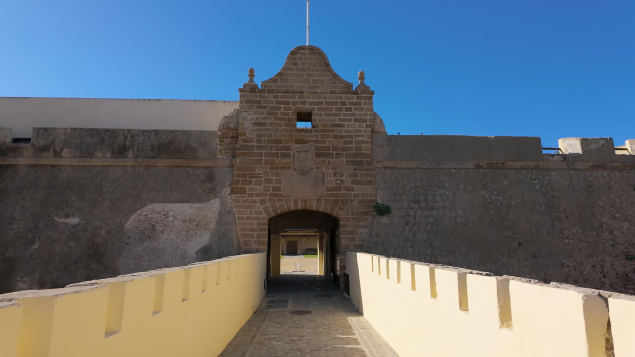 A stone gateway at the entrance of an ancient fortress in C&aacute;diz, with a path leading through it, flanked by high walls under a clear blue sky