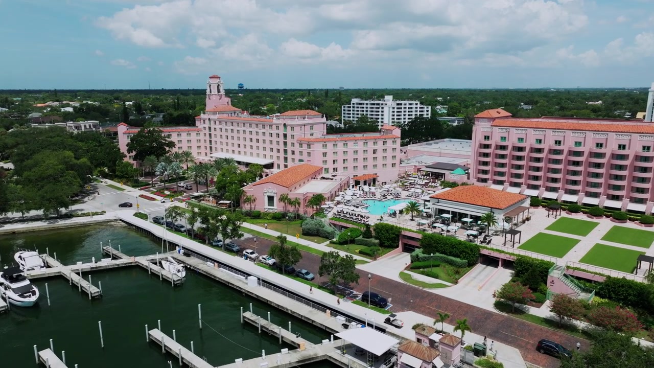 Vista aerea dell'iconico Vinoy Hotel rosa e della sua marina nel centro di St. Petersburg, Florida, con yacht di lusso ormeggiati lungo il molo sotto un cielo estivo luminoso.