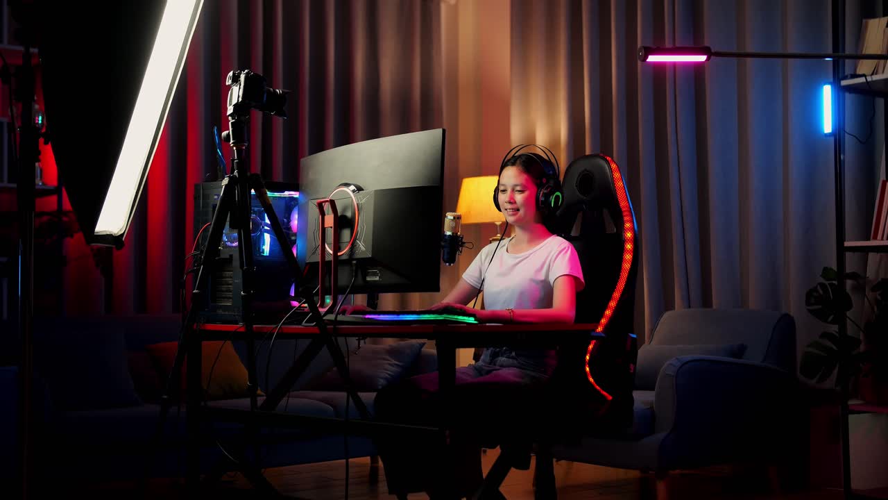 chica asiática sonriendo a la cámara mientras juega un juego a través de la red en una computadora personal. videojuego de transmisión en vivo, escritorio iluminado por luz de tira led rgb
