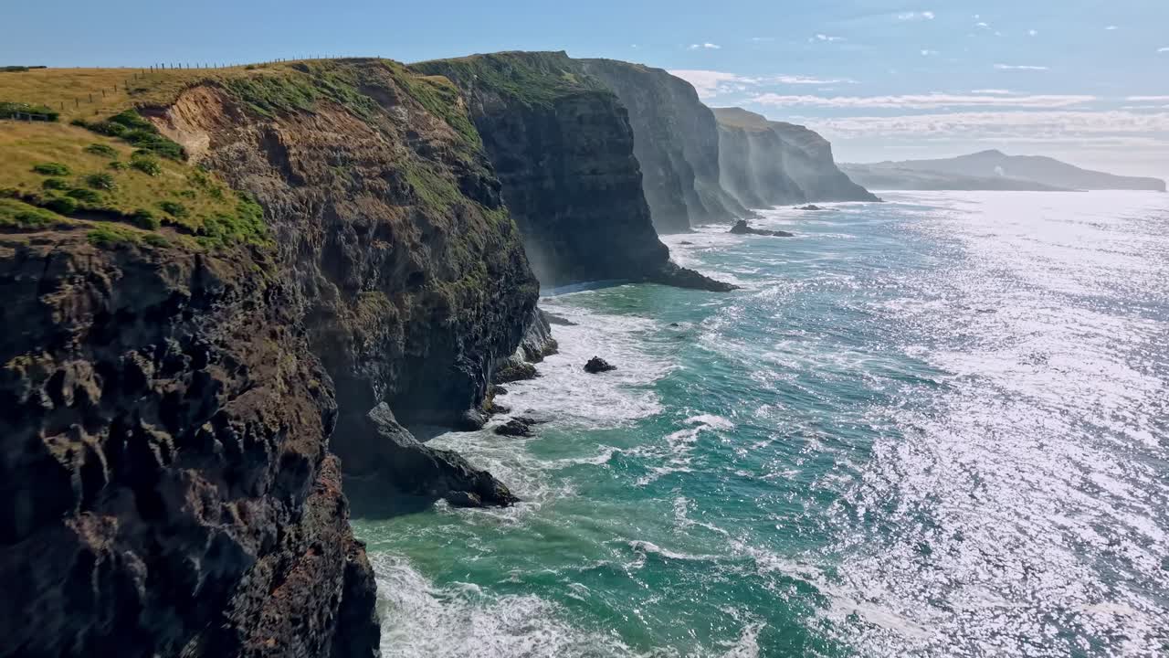 Drone shot flying forward along the volcanic cliffs of Māori Head on the Otago Peninsula near Dunedin, New Zealand, showing waves crashing on the rocks below