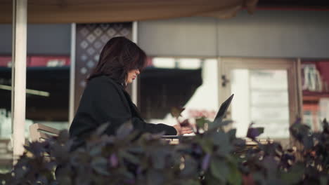 freelancer trabaja en una computadora portátil en un restaurante al aire libre, vista lateral, con un elegante abrigo negro con flores decorativas, reflejando la vida urbana en edificios de vidrio cercanos