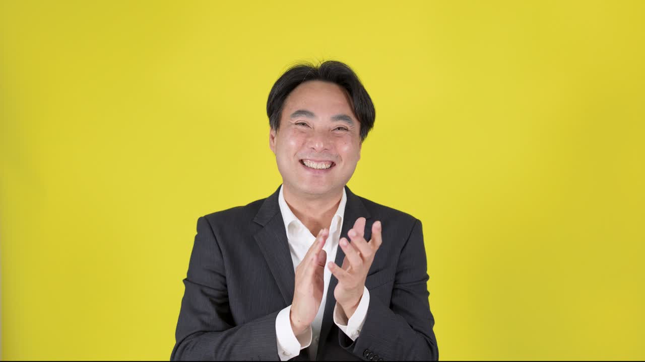 自信に満ちたビジネスマンが拍手をして、明るいスタジオ照明の中で笑顔で親指を立てています。カメラは固定されています。