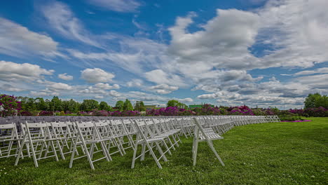 static shot of rows of white wooden folding chairs on the green grass surrounded by floral garden with white clouds passing by in timeplase