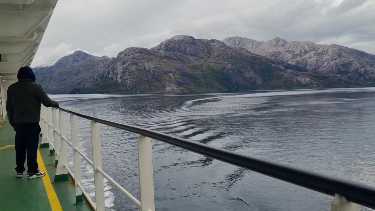 Männlicher Reisender geht entlang der Fährreling auf einer Reise durch die Fjorde des chilenischen Patagoniens, umgeben von dramatischen Bergen und ruhigem Wasser während einer abenteuerlichen Panoramafahrt.