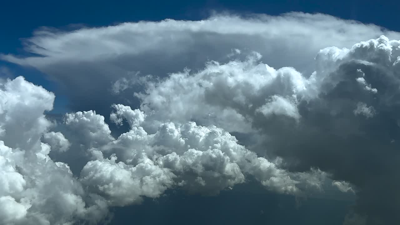 an aerial view taken from a jet cockpit flying at cruise through a sky full of storm clouds, and a anvil shape cumulonimbus ahead, under a blue sky. Ultra-realistic 4K.