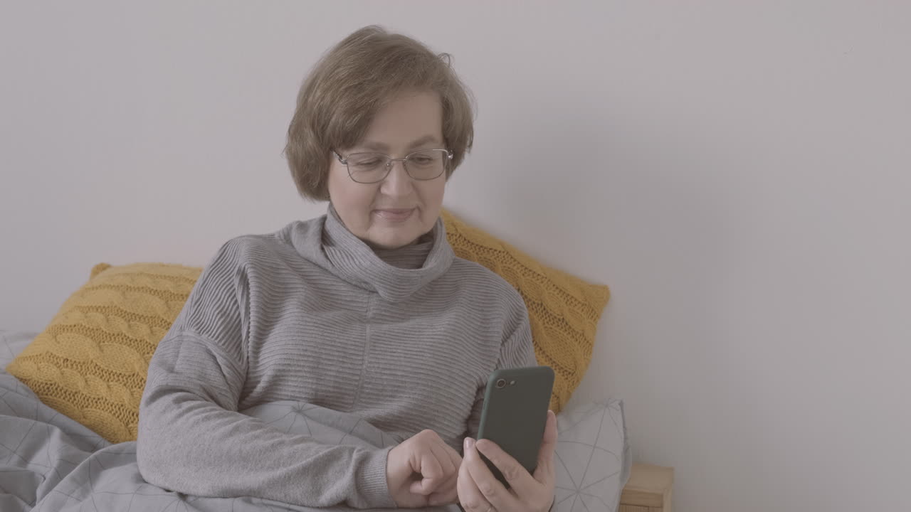 Senior Woman With Eyeglasses Sitting On Bed Making A Video Call On A Smartphone