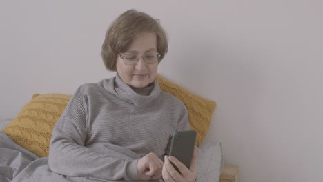 senior woman with eyeglasses sitting on bed making a video call on a smartphone