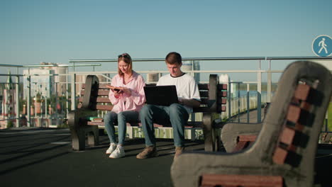 male and female students studying outdoors on a bench; male typing on laptop while engaging with female partner reading from her book, set against an urban skyline backdrop