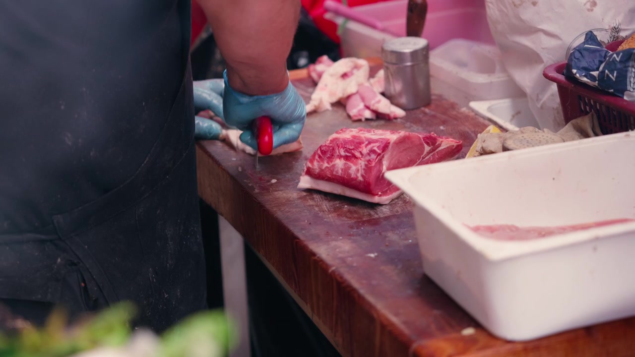 Um açougueiro usando luvas corta pedaços frescos de carne em um bloco de madeira em uma barraca de mercado siciliano tradicional, em plano médio.