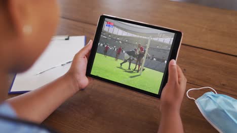 Composite-of-woman-sitting-at-table-with-face-mask,-watching-rugby-match-on-tablet