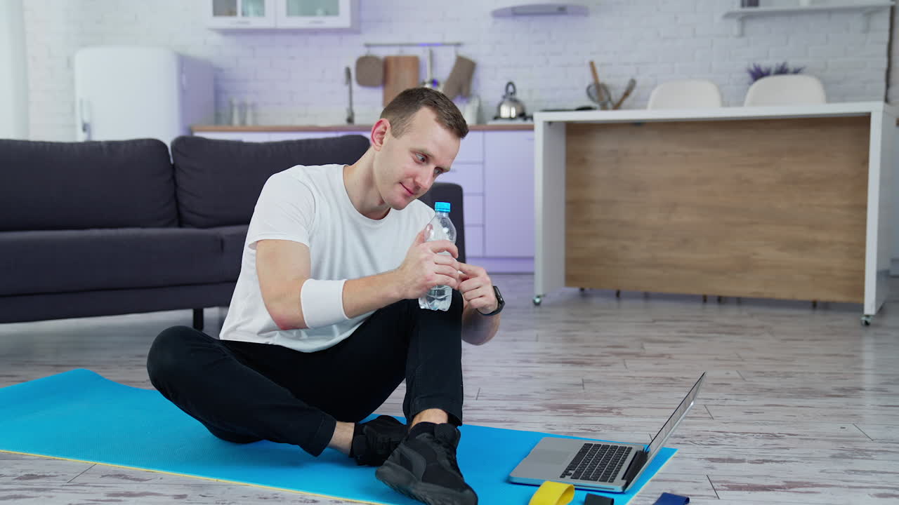 Young man sitting on a mat after training in front of a laptop. Sportive guy talking to someone through the laptop and shows a bottle of water in the kitchen.