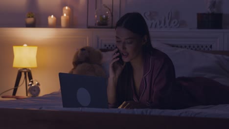 woman on bed talking on the phone and using a laptop at night