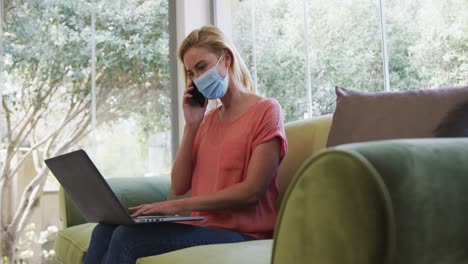 woman wearing face mask using laptop talking on smartphone at home