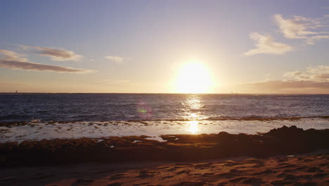 Von-Einem-Sandstrand-Auf-Diamond-Head,-Hawaii,-Oahu,-Geht-Eine-Strahlend-Weiße-Sonne-Hinter-Dem-Pazifischen-Ozean-Unter,-Während-Die-Wellen-Des-Pazifiks-Auf-Den-Sandstrand-Rollen