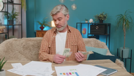 mature man at table with documents bills holding money cash showing financial distress at home