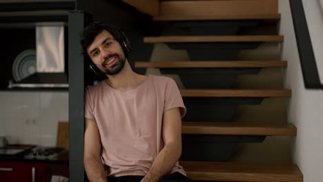 Hombre-Barbudo-Sonriente-Con-Auriculares,-Sentado-En-Escaleras-De-Madera-En-Casa-En-Cuadros