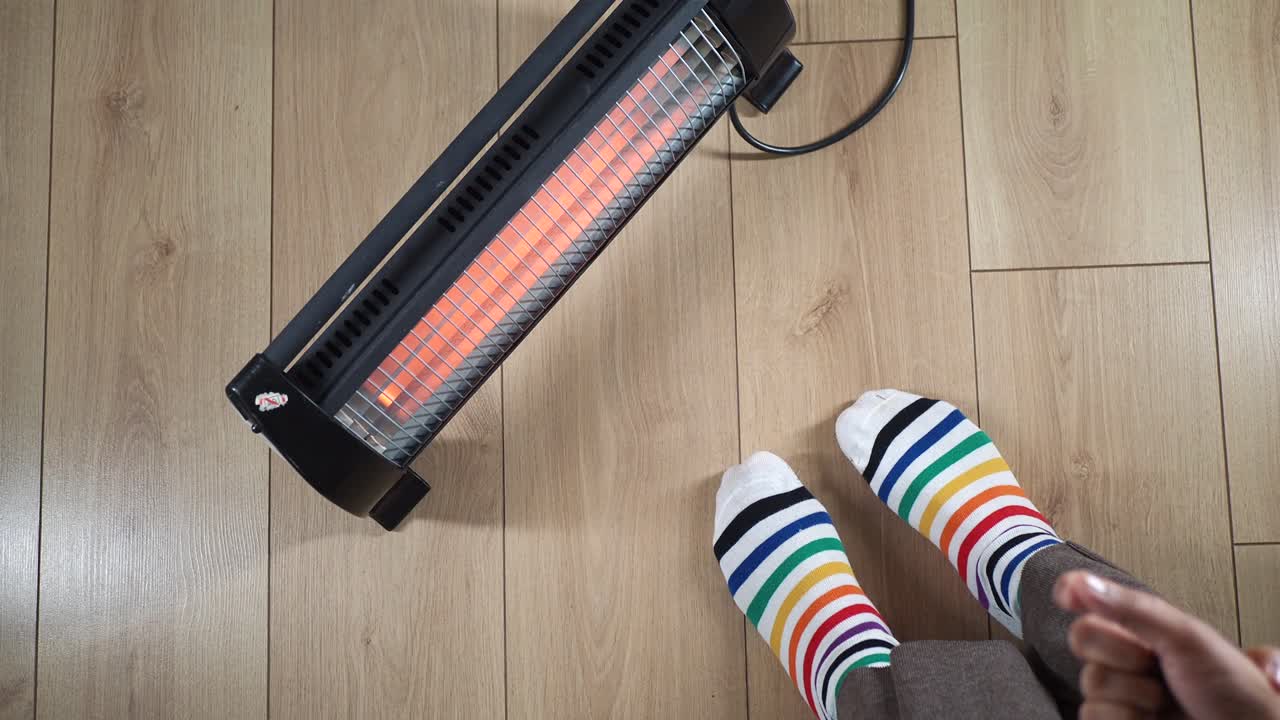 Person Warming Feet and Hands with Electric Heater on Wooden Floor