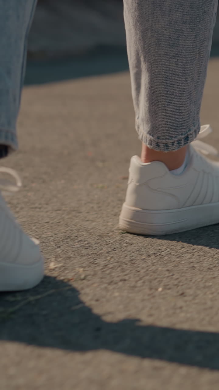 vista trasera de primer plano de un individuo caminando por un camino asfalto iluminado por el sol, con zapatillas blancas y vaqueros, las sombras se proyectan en el suelo, capturando el movimiento casual, el entorno al aire libre