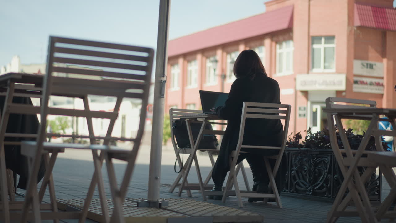 vista trasera de una mujer profesional escribiendo en una computadora portátil al aire libre en el centro comercial, vista parcial de alguien con un traje azul caminando en la distancia, rodeado de sillas de madera y plantas en maceta