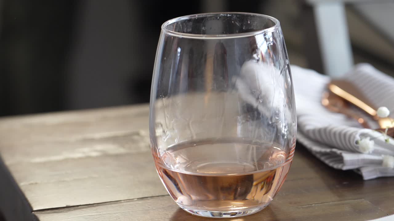 Woman Picks Up Stemless Wine Glass of Rose Half-Full from Wooden Table, Closeup of Hand Wearing Pink Nail Polish and Kitchen Table Napkin and Silverware in Background, White and Golden Red Color