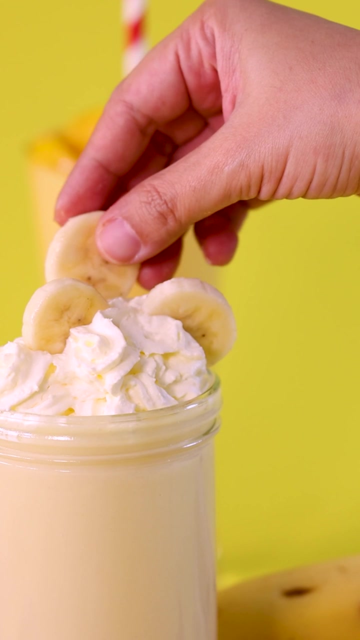 A hand carefully garnishes a creamy banana milkshake topped with whipped cream against yellow.