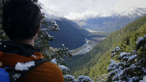 Mann,-Der-Auf-Einem-Abenteuer-Durch-Schneebedeckte-Berge-Wandert-Und-Auf-Ein-Wunderschönes-Grünes-Tal-Blickt,-Durch-Das-Ein-Fluss-Fließt