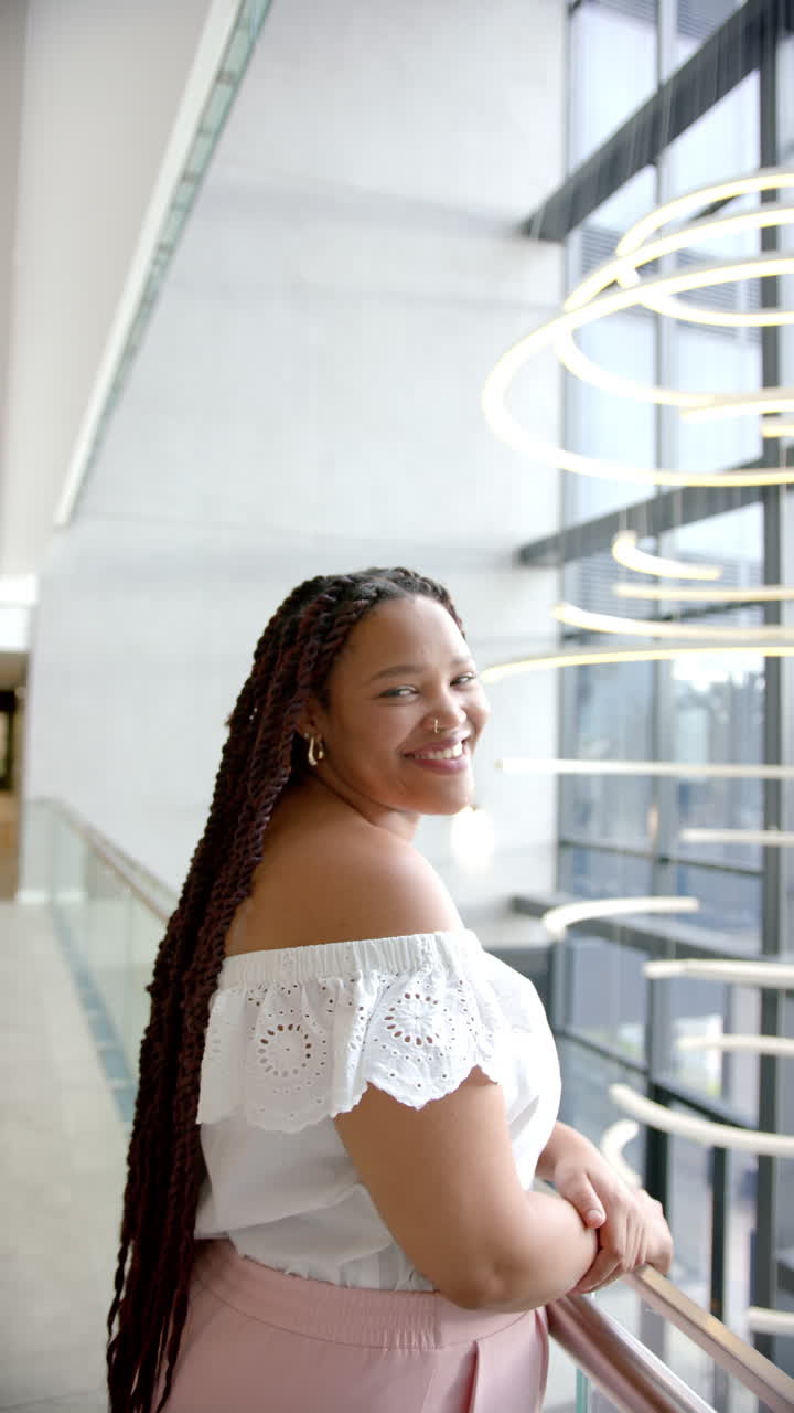 Vertical video: Smiling woman with long braids standing by modern window railing indoors