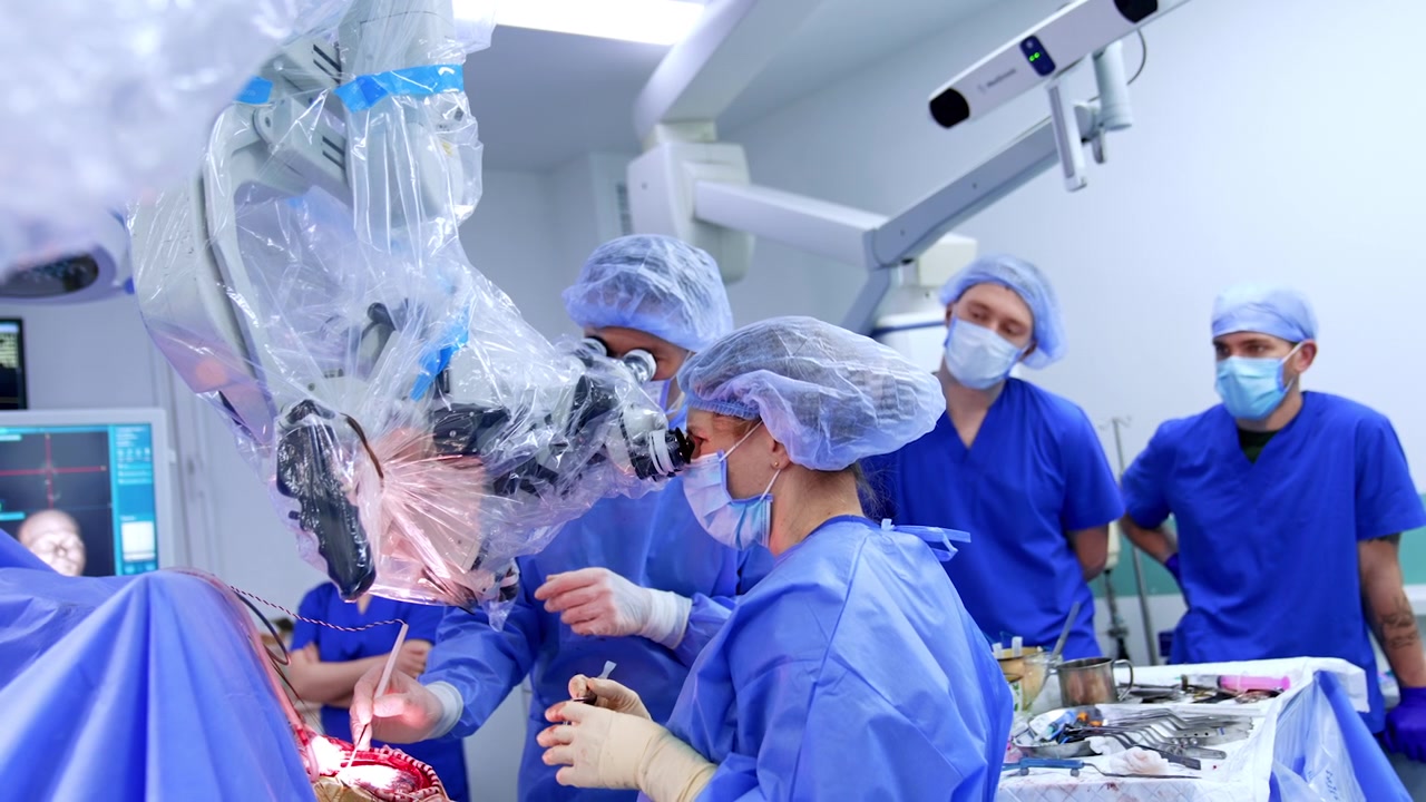 Surgical team with neuronavigation monitors performing open brain surgery. Multiple surgeons in blue gowns, two monitors show brain MRI, instrument tray at foreground.