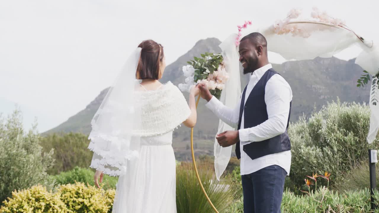 Video of happy diverse bride and groom holding hands, smiling and dancing at outdoor wedding