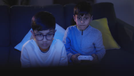 Vista-Frontal-De-Dos-Niños-Pequeños-En-Casa-Divirtiéndose-Jugando-Con-Una-Consola-De-Juegos-De-Computadora-En-La-Televisión-Sosteniendo-Controladores-A-Altas-Horas-De-La-Noche-1