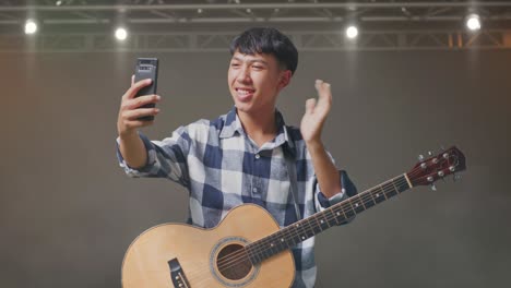 adolescente asiático con la mano agitando la guitarra y haciendo una llamada de video en el teléfono inteligente mientras está de pie en el escenario