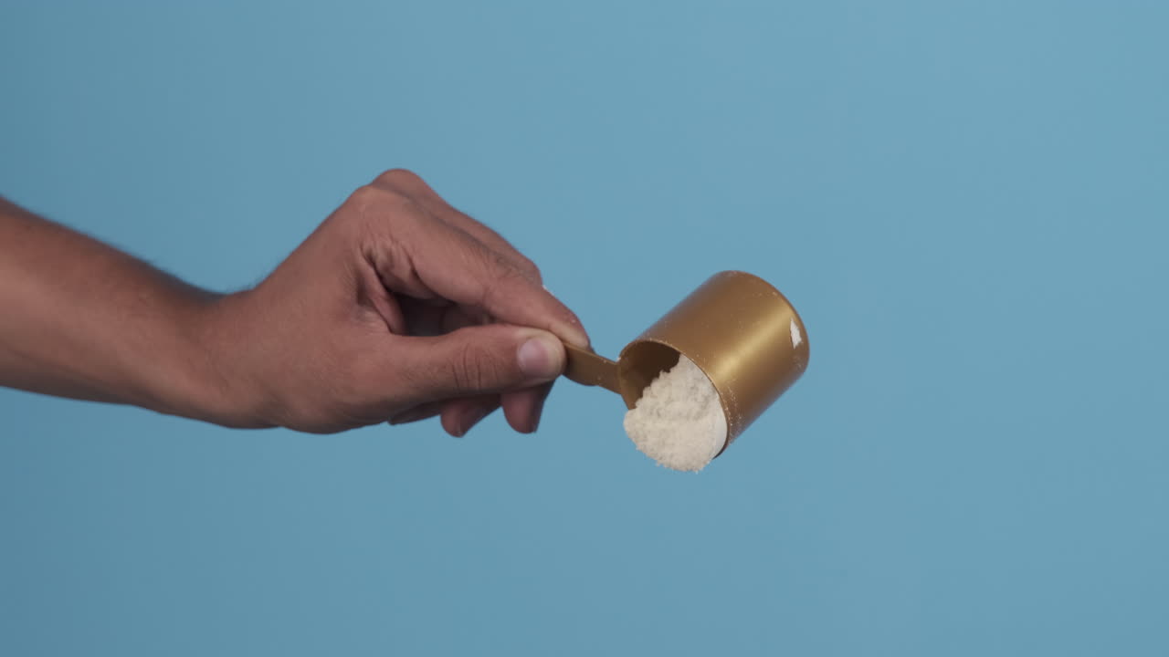 A hand holding and measuring protein powder with a scoop against a blue background