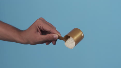 a hand holding and measuring protein powder with a scoop against a blue background