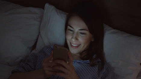 close-up view of the charming caucasian woman with dark hair lying in the bed, chatting on the smartphone late at night