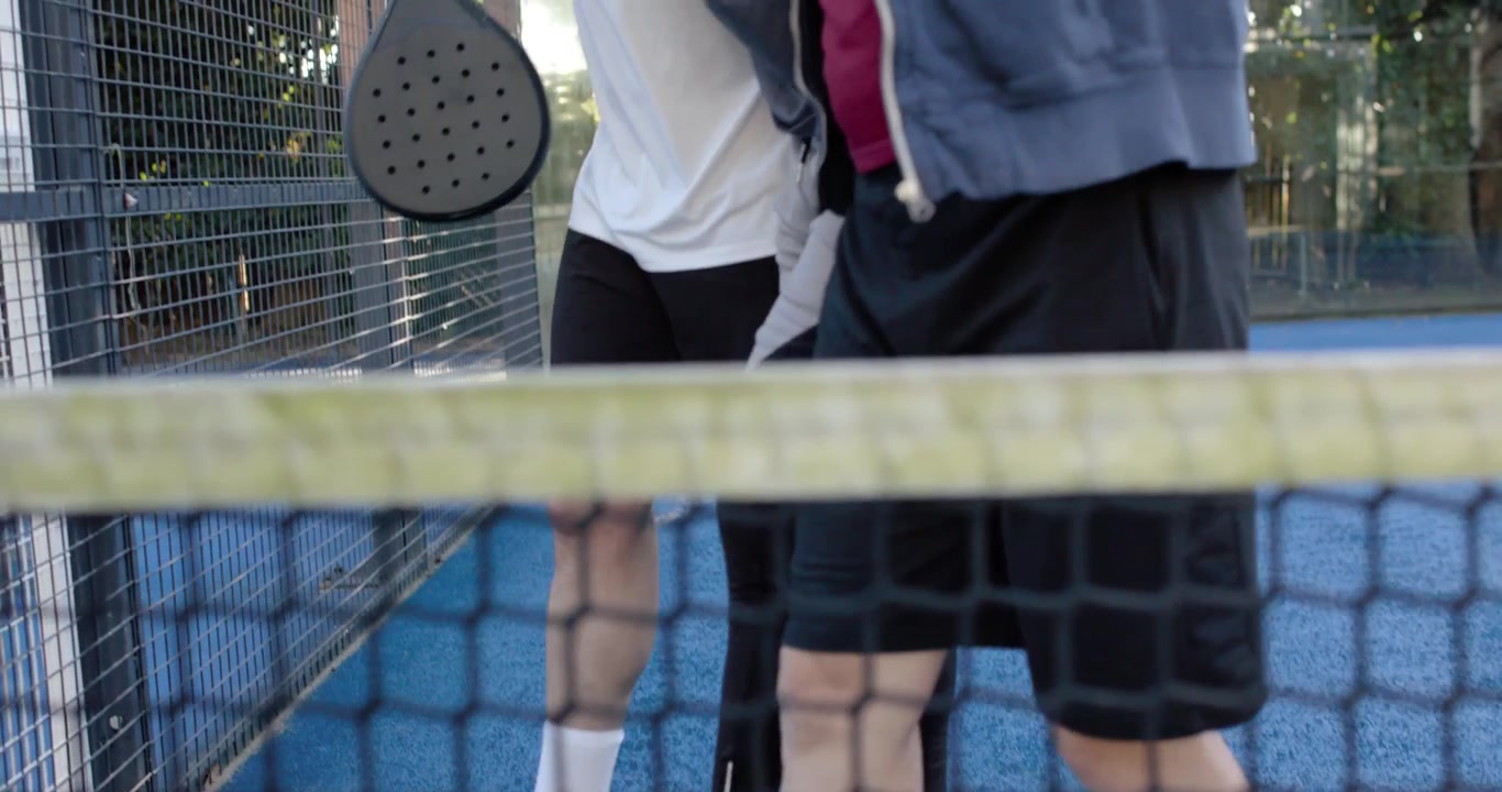 Diverse men shifting feet stepping past net holding black padel racket exiting court with balls