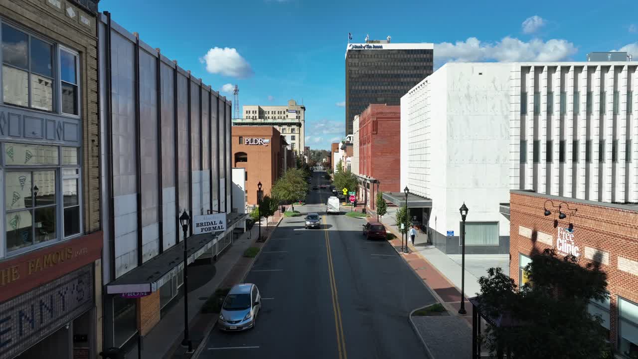 Aerial establishing shot of parking cars on main street of center in sunlight. Stores and Markets in old buildings. Walking pedestrian on sidewalk. Forward flight.