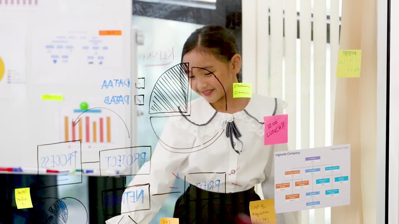 Una niña con blusa blanca dibuja gráficos de negocios en un cristal en una oficina luminosa.