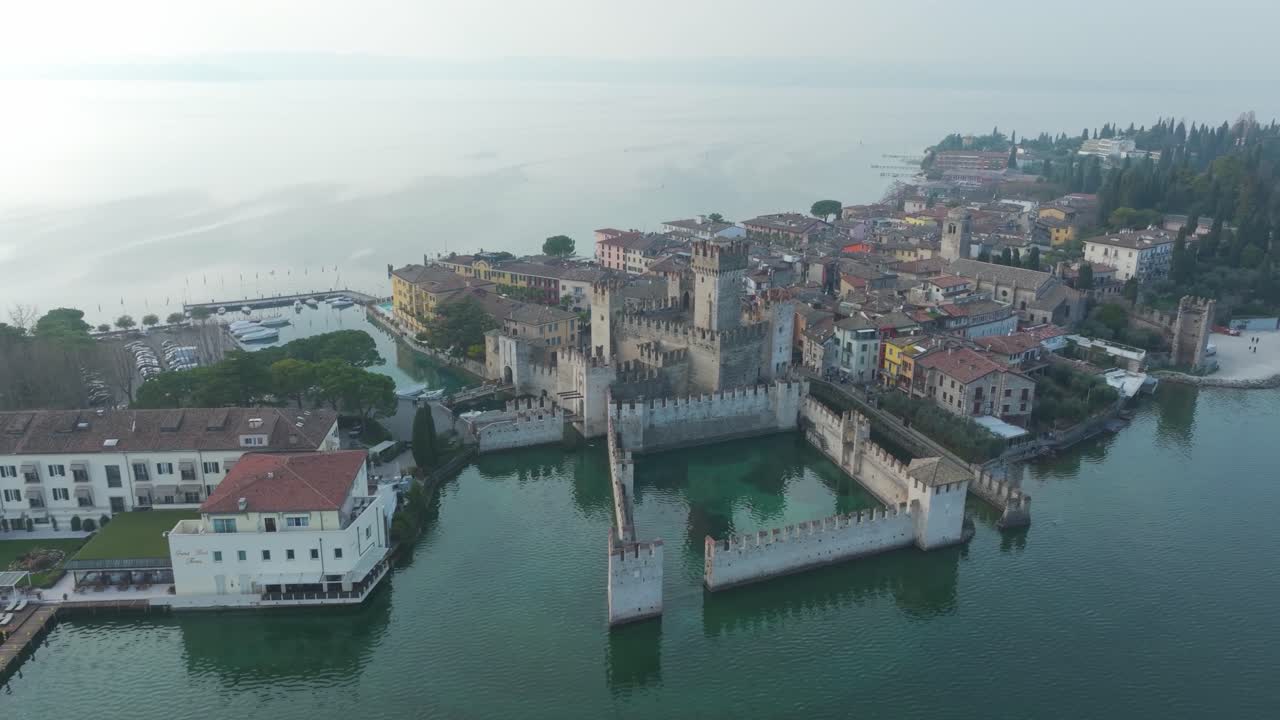 Drone sobrevoando o Castelo Scaligero em Sirmione, Itália, revelando as águas turquesa do Lago de Garda, casas coloridas e muros históricos em um lindo dia, retroiluminado.