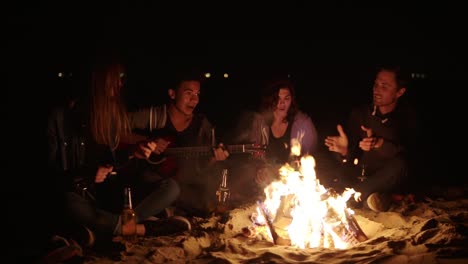 Late-night-picnic-of-young-people-with-bonfire-in-the-evening.-Cheerful-friends-singing-songs-and-playing-guitar,-talking-and