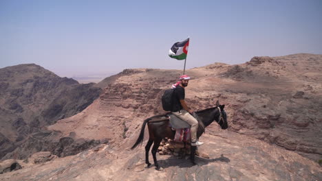 man on mule by jordanian national flag on lookout above petra archaeological site and canyon, full frame, pan