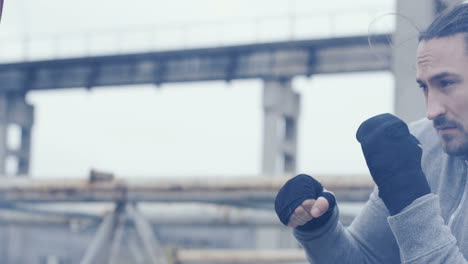 caucasian man with long hair hitting a punching bag outdoors an abandoned factory on a cloudy morning