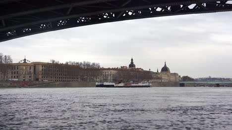 Ein-Frachtschiff-Gleitet-Entlang-Der-Ruhigen-Rhone,-Unter-Der-Gewölbten-Stahlbrücke-Von-Lyon,-Inmitten-Alter-Europäischer-Stadtlandschaften