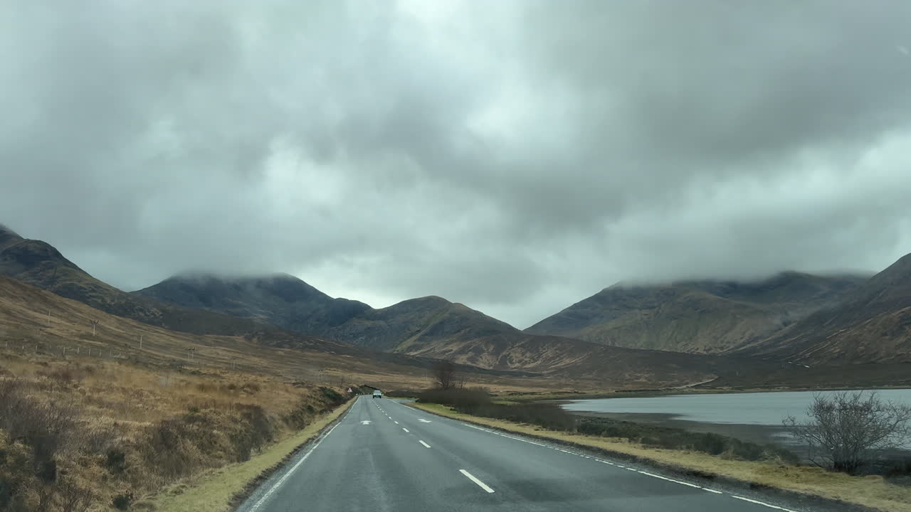 A car drives through the moody Scottish Highlands on an overcast day, with cloud-covered hilltops creating a serene, misty landscape full of wild, untamed beauty and quiet solitude.