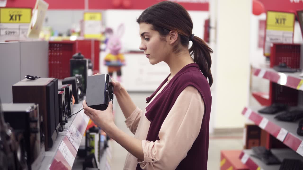 una joven mujer que elige un sistema de audio de una variedad en una tienda de electrónica. examinando cuidadosamente los altavoces en una fila de vitrinas.