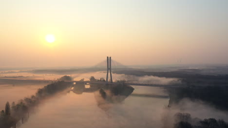 Aerial-shot-over-a-wide,-calm-river,-during-a-foggy-morning,-close-to-a-modern-cable-road-bridge