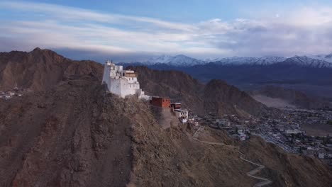 Toma-Cinematográfica-De-Un-Dron-Que-Revela-El-Monasterio-Y-El-Templo-De-Sankar-Con-Vistas-A-La-Ciudad-De-Leh-Con-La-Vista-De-La-Cordillera-Nevada-Del-Himalaya-Al-Fondo-Durante-La-Puesta-De-Sol,-Ladakh,-India