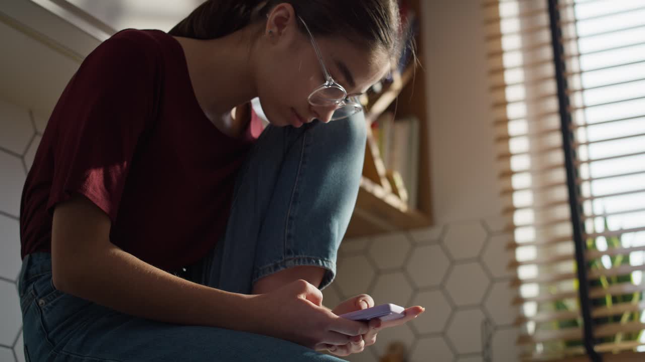 Low angle view of caucasian teenager girl sitting in the kitchen and using mobile phone in silence